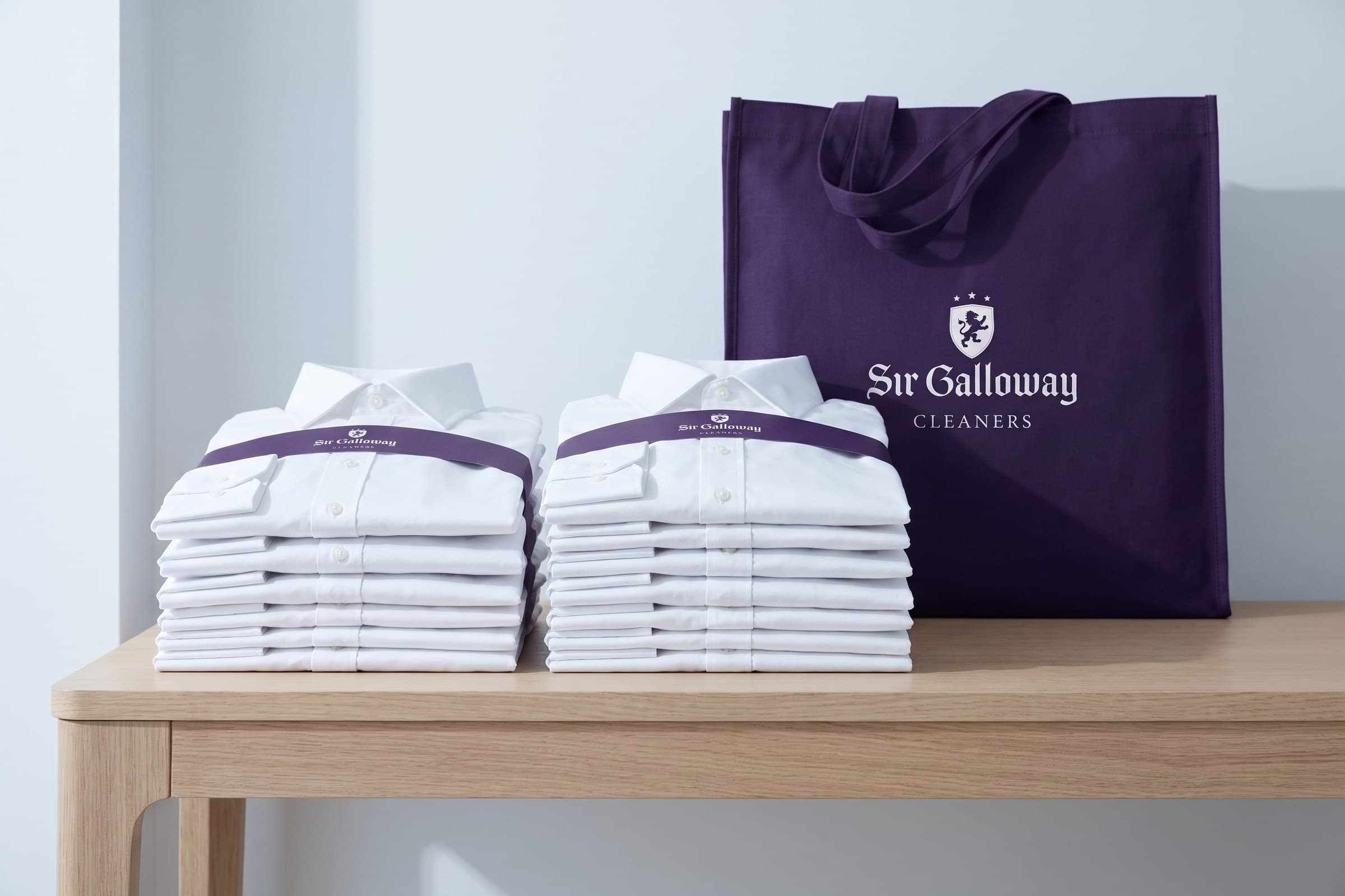A canvas tote of folded white linens banded in plum, set on a console table by a window.