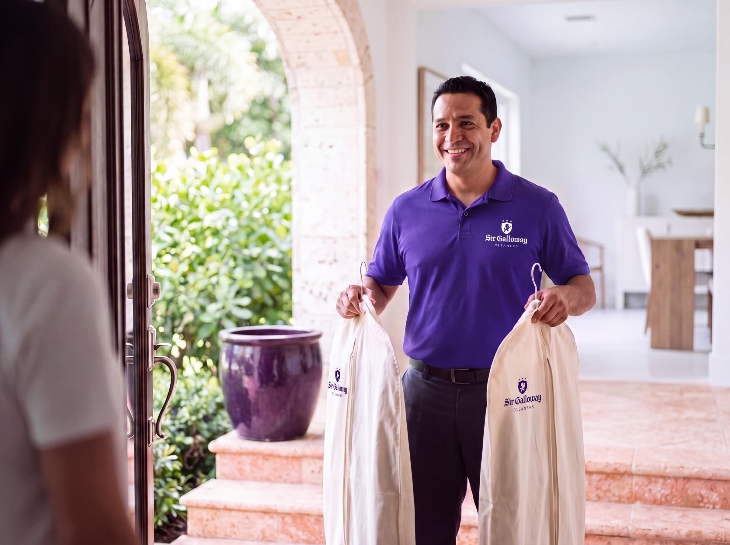 A Sir Galloway courier in a purple polo smiling as he hands two crested Sir Galloway garment bags to a client at her front door.