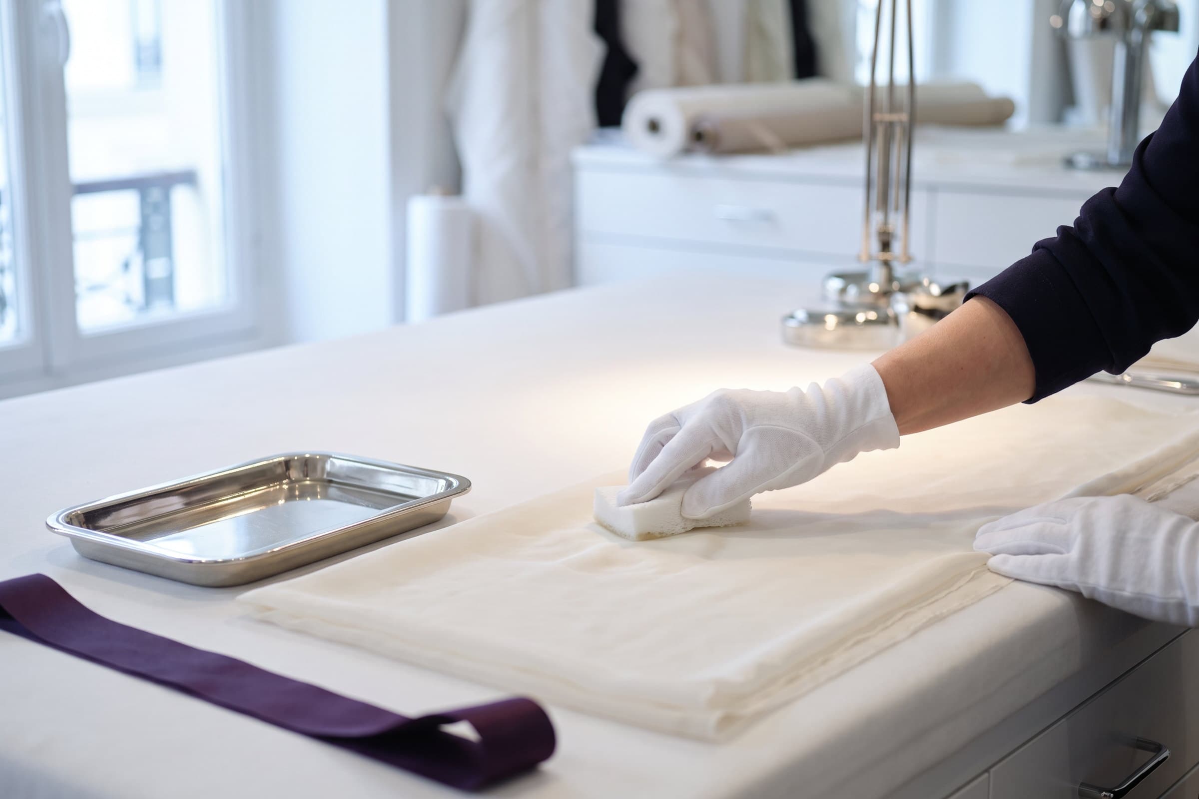 Gloved hands working a small sponge over folded ivory fabric on a padded white worktable, a steel tray and a plum ribbon alongside.