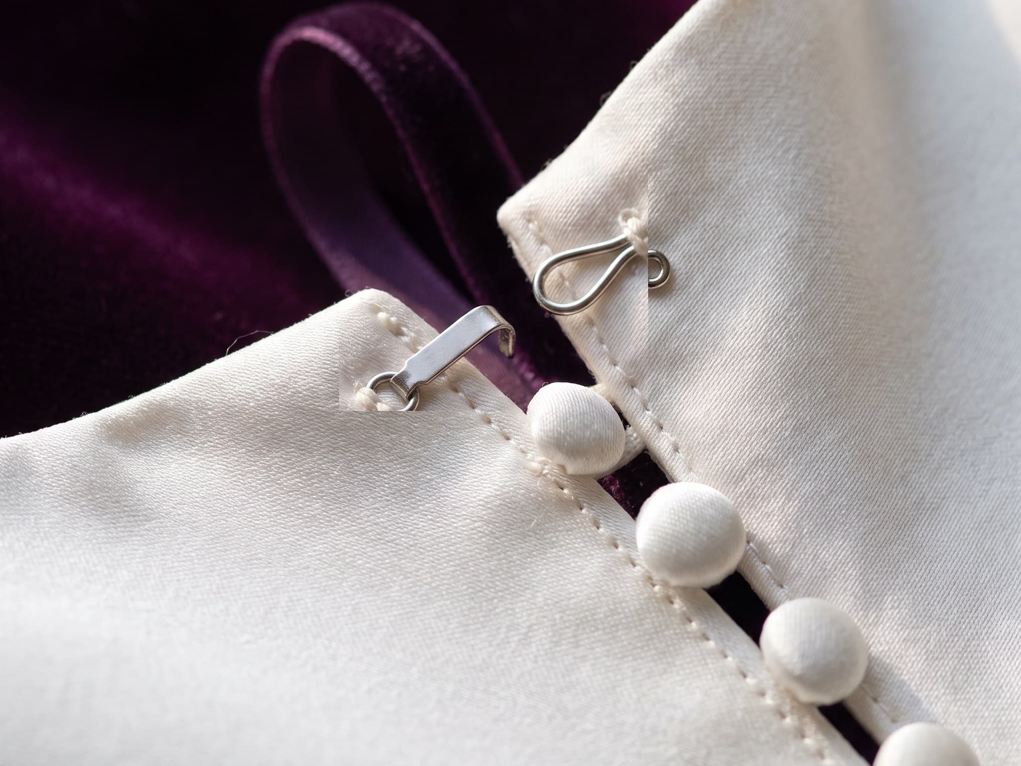 Close-up of a hook and bar closure and covered satin buttons at the back of an ivory wedding gown, on deep purple velvet.