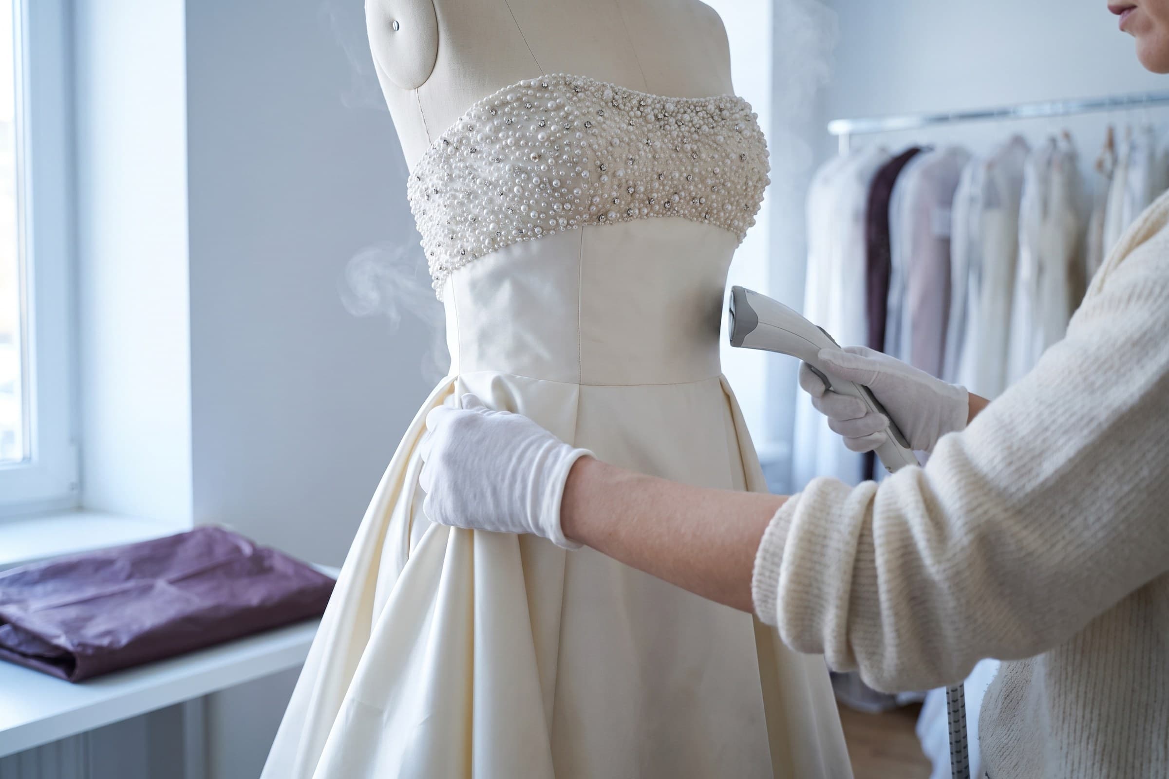 An ivory off-the-shoulder wedding gown on a dress form in a bright studio, with a folded garment bag on a bench beside it.