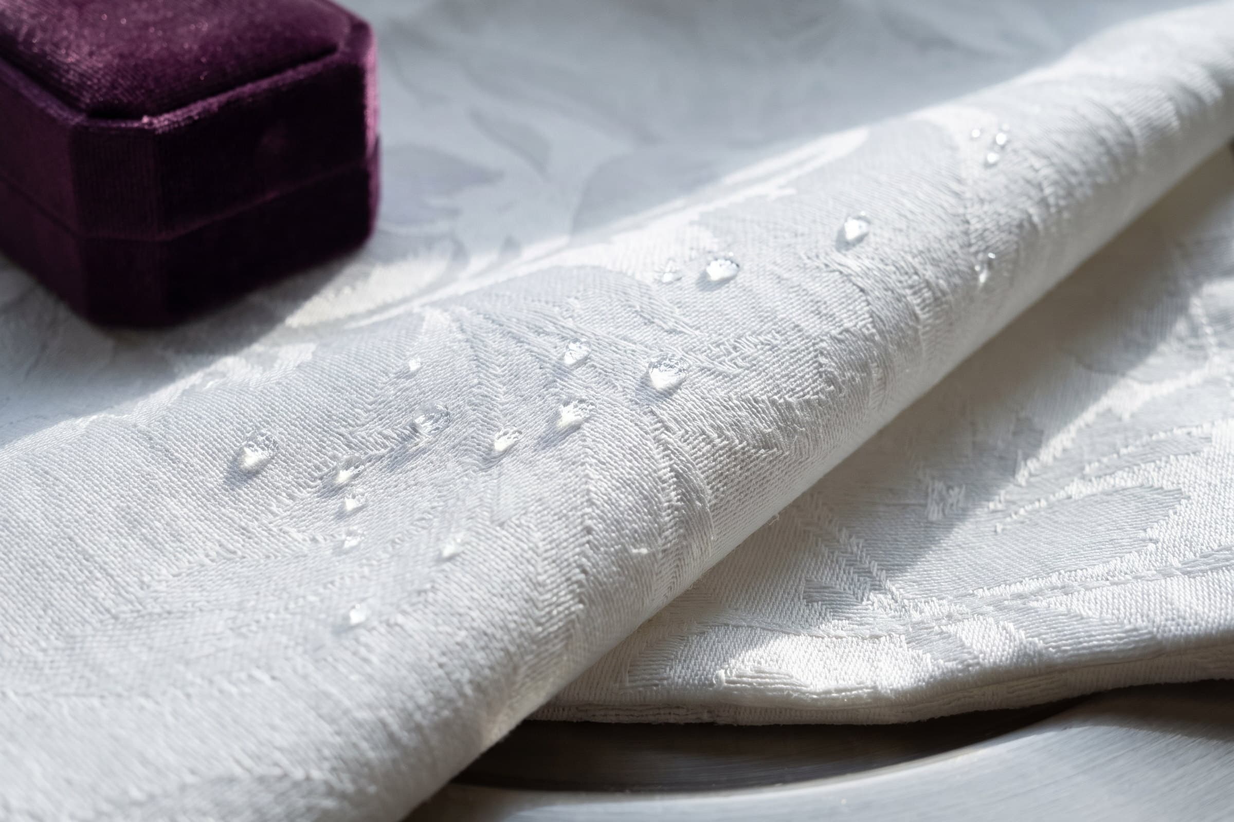 Water beading on the surface of a white damask linen, with a plum velvet box beside it.