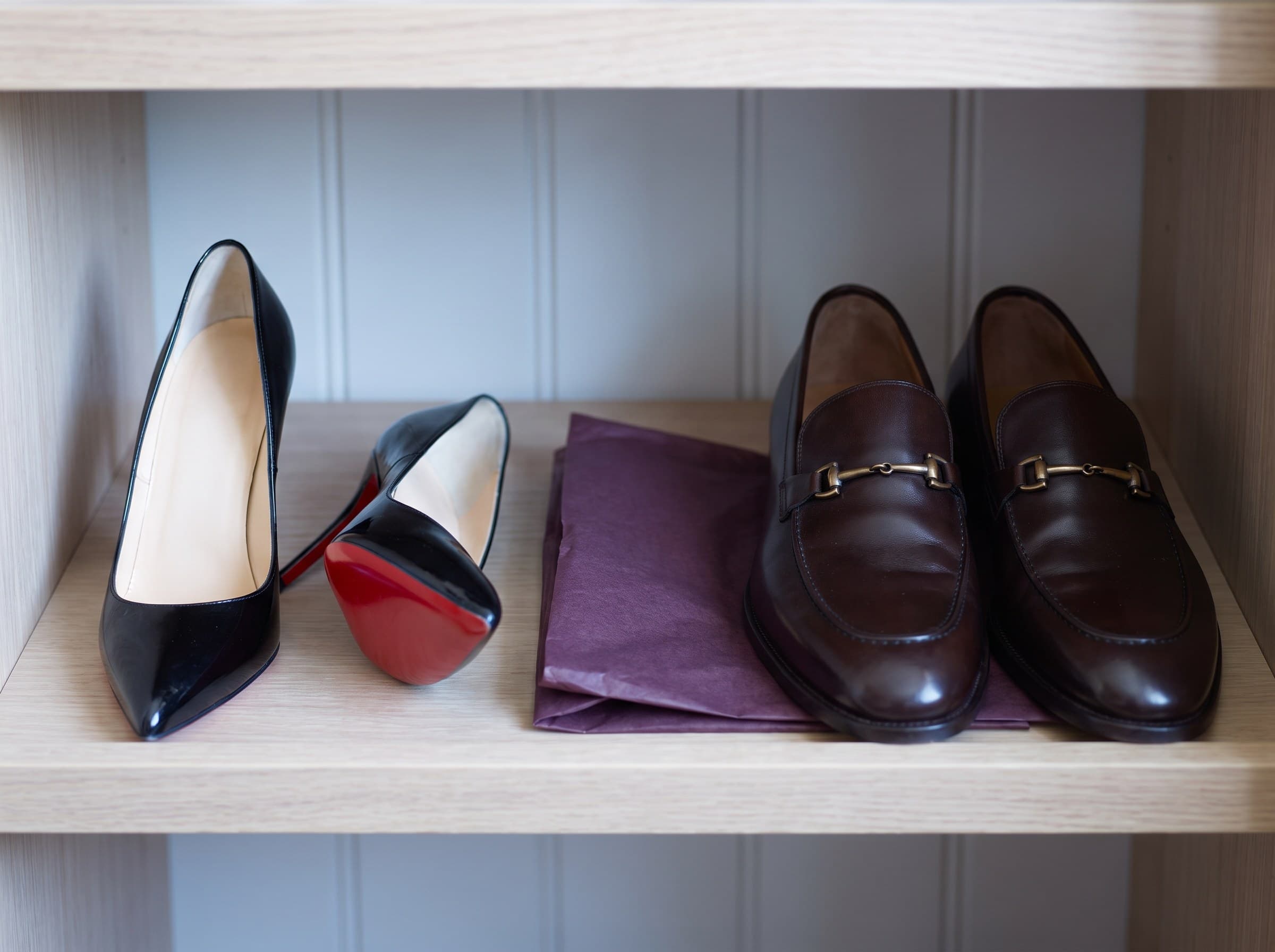 A pair of polished black patent pumps, one on its side showing the red lacquered sole, beside plum tissue paper.