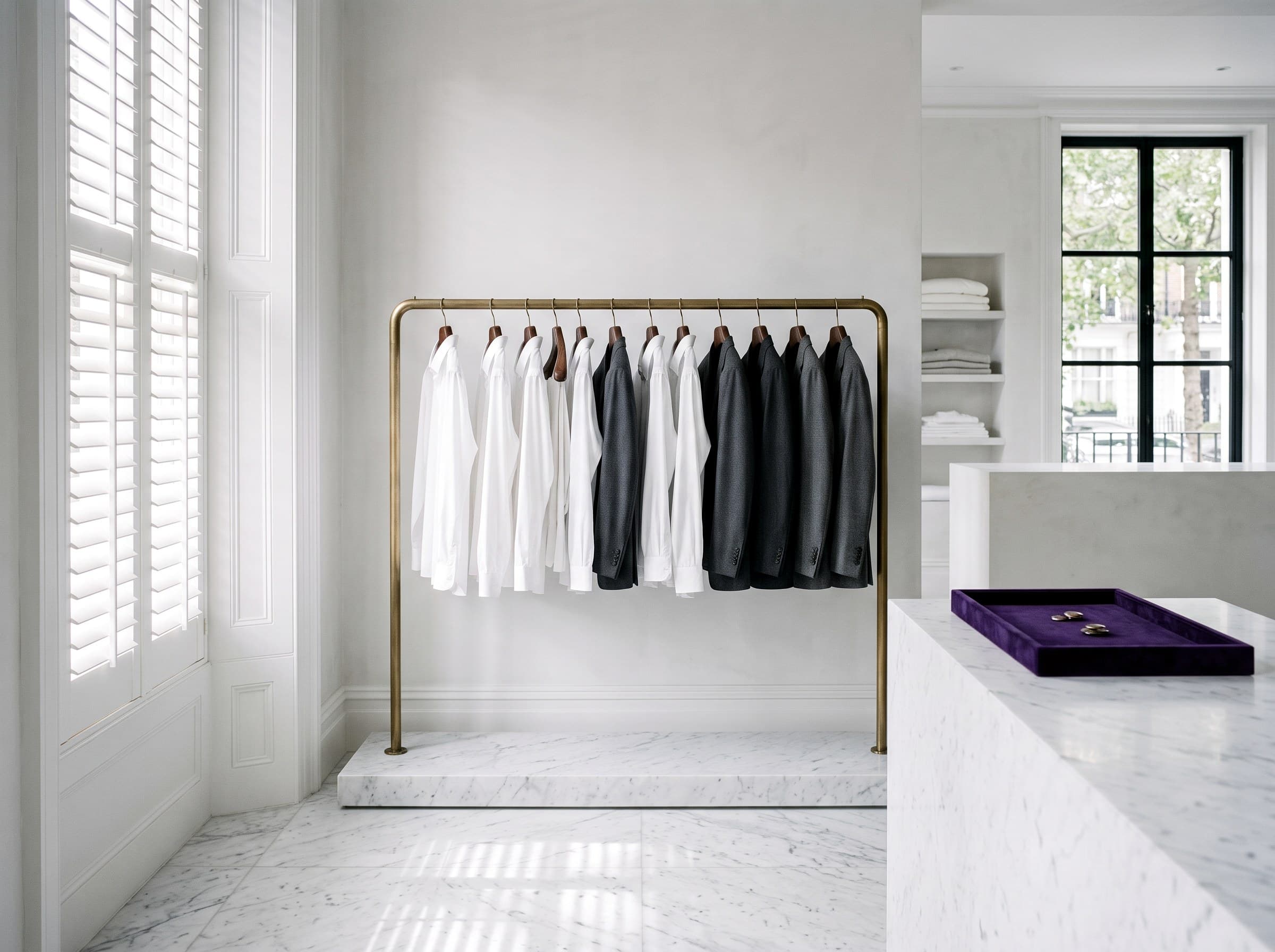 A wide editorial view of the Sir Galloway workroom, brass rail with white shirts and charcoal jackets, plantation shutter light, polished marble floor.