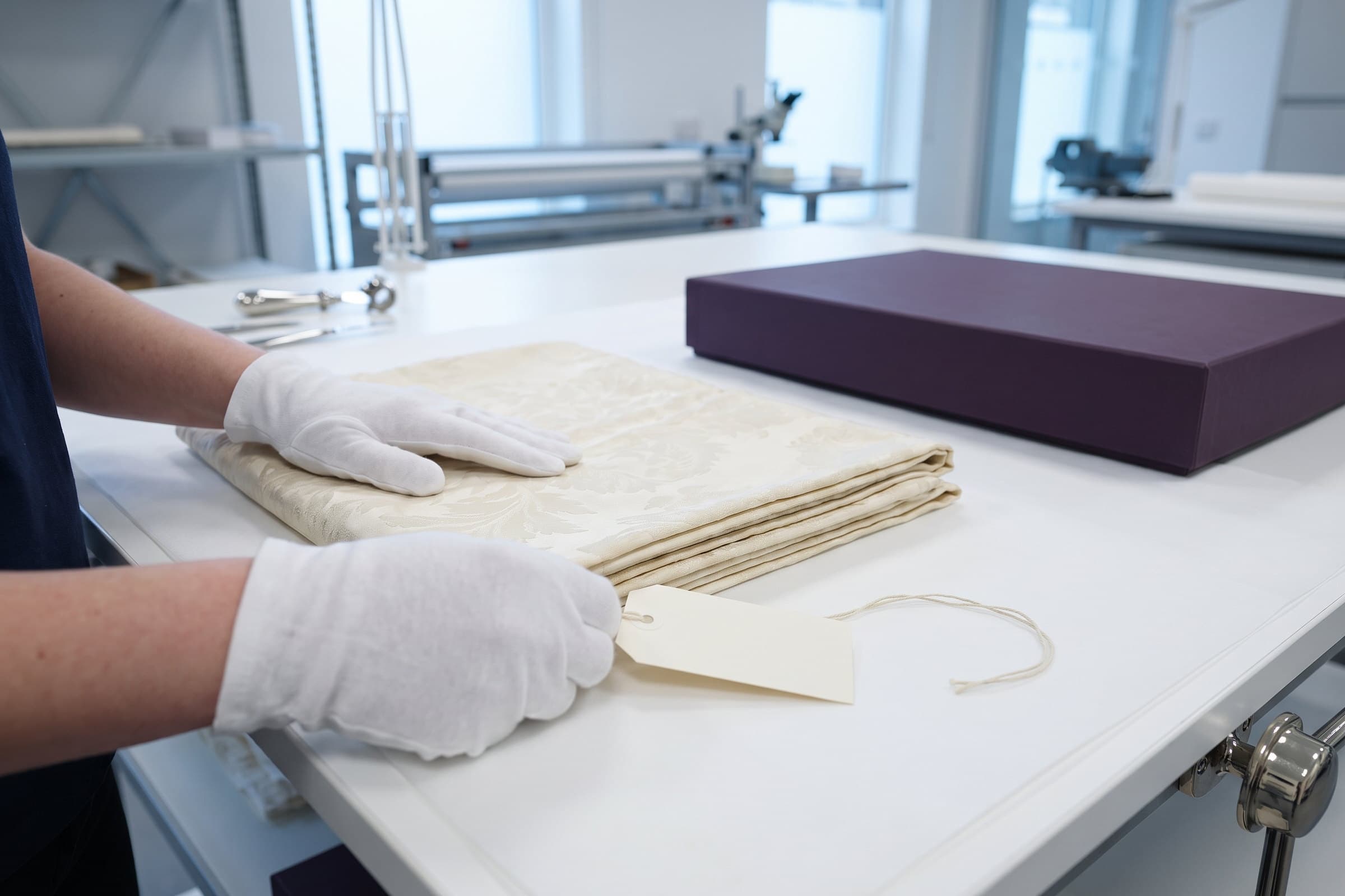 Gloved hands smoothing a folded cream damask linen on a worktable beside a blank string tag.