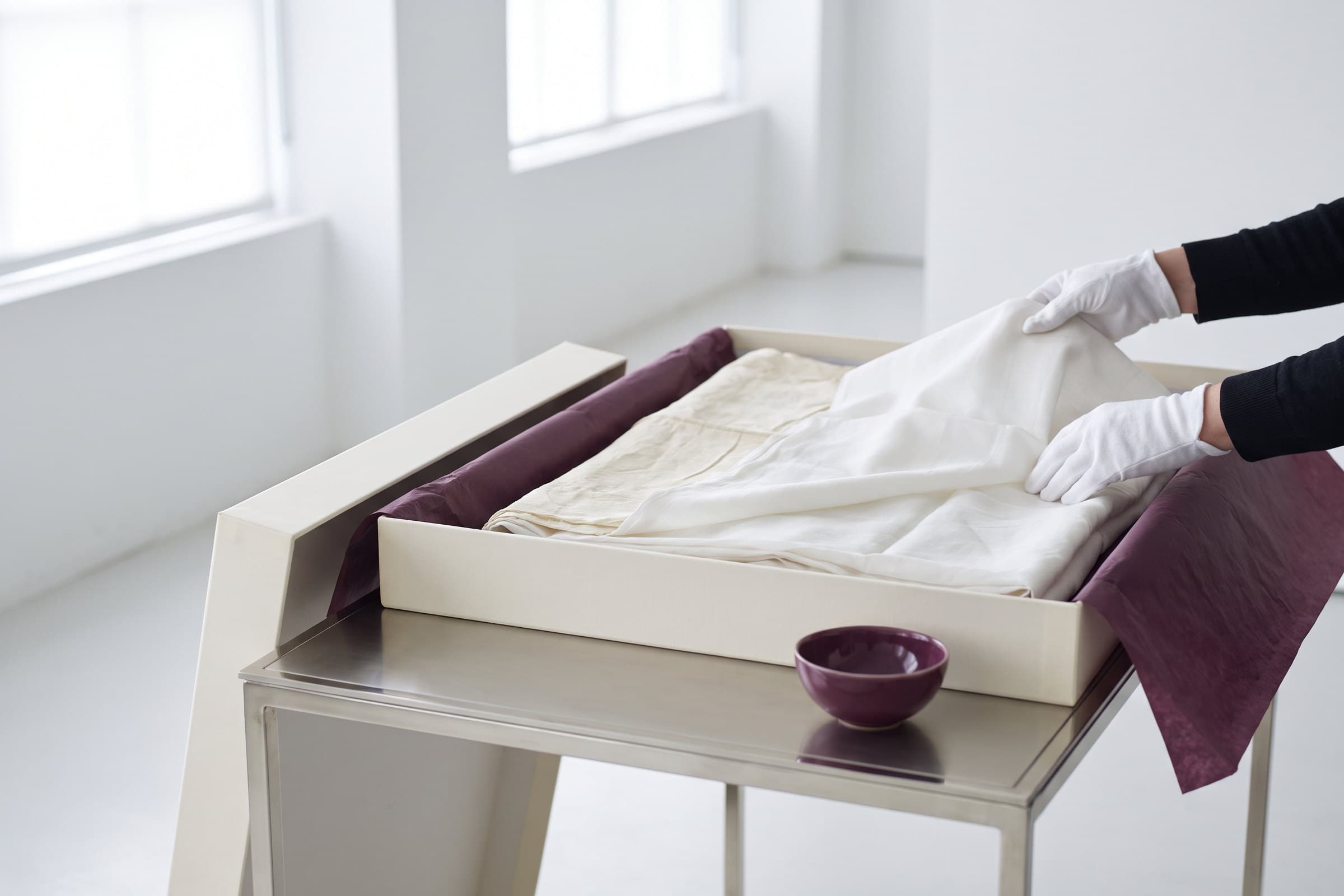 Gloved hands laying folded cream textiles into an archival box lined with plum tissue.
